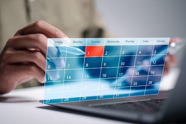 Ein Kalender, der einen Tag im Dienstag hervorgehoben zeigt, wird von einer Hand gehalten, während ein Laptop im Hintergrund sichtbar ist.